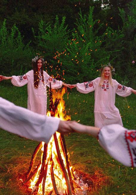 Kobiety w białych sukniach tańczące wokół ognia na polanie.